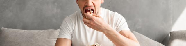 Single man 20s sitting on sofa in gray apartment and eating pop corn, while watching tv in morning