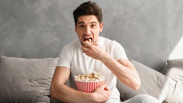 Single man 20s sitting on sofa in gray apartment and eating pop corn, while watching tv in morning