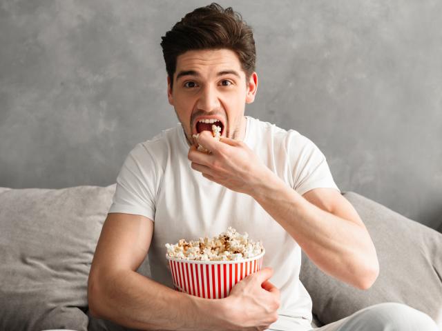 Single man 20s sitting on sofa in gray apartment and eating pop corn, while watching tv in morning