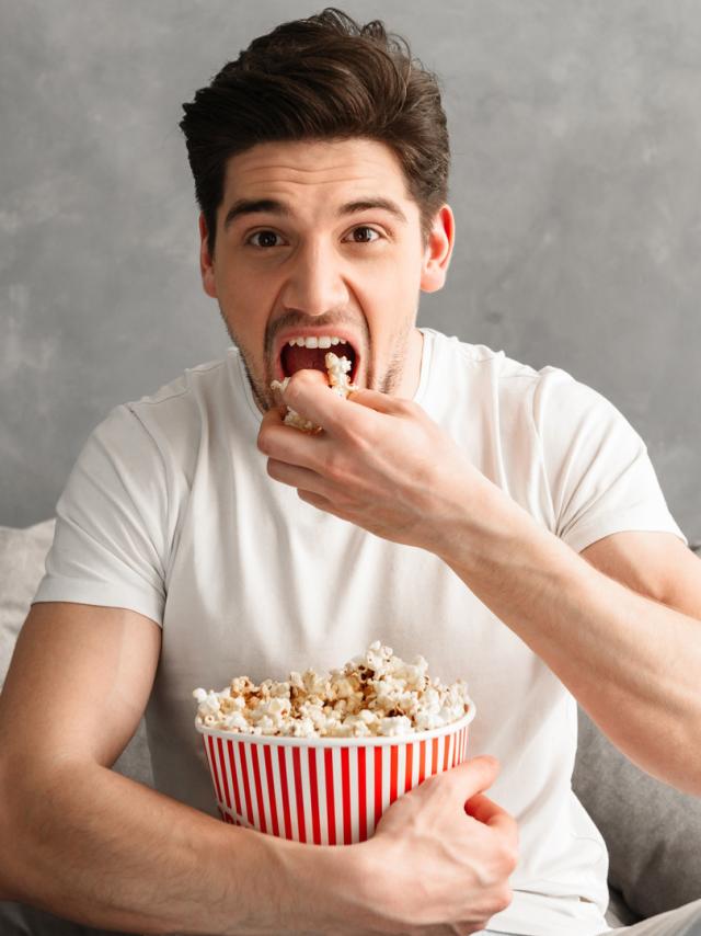 Single man 20s sitting on sofa in gray apartment and eating pop corn, while watching tv in morning