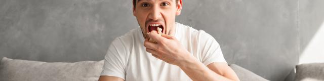 Single man 20s sitting on sofa in gray apartment and eating pop corn, while watching tv in morning