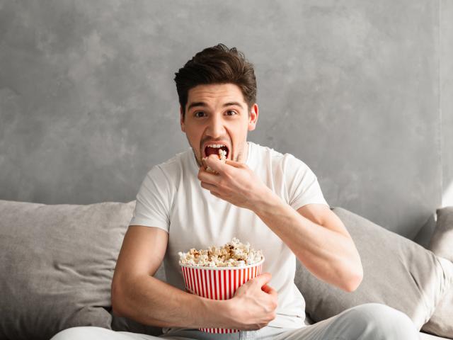 Single man 20s sitting on sofa in gray apartment and eating pop corn, while watching tv in morning