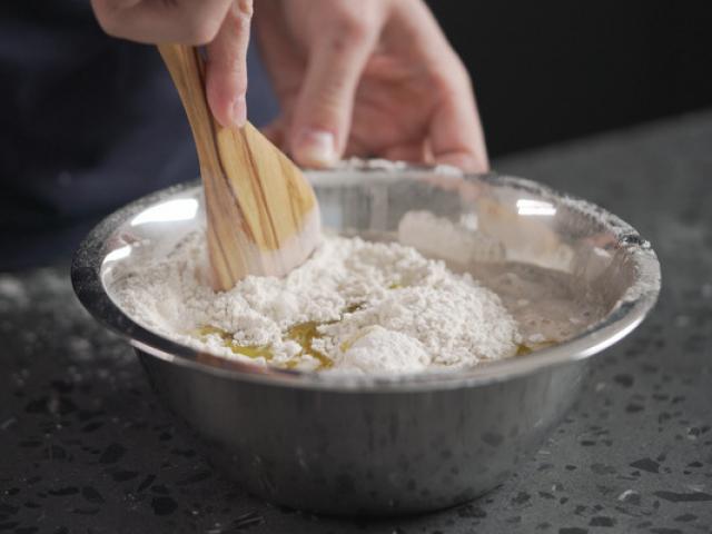 man mixing wet ingredients into flour in steel bowl on concrete countertop