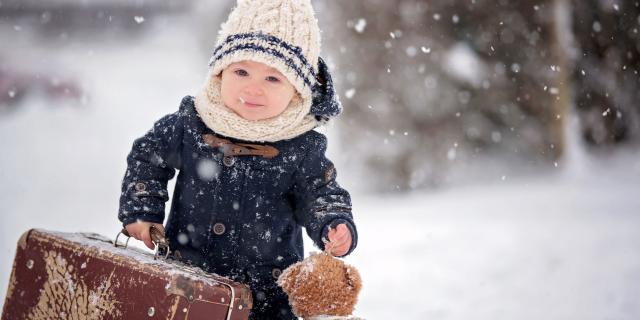 Baby playing with teddy in the snow, winter time. Little toddler boy in blue coat, holding suitcase and teddy bear, playing outdoors in winter park