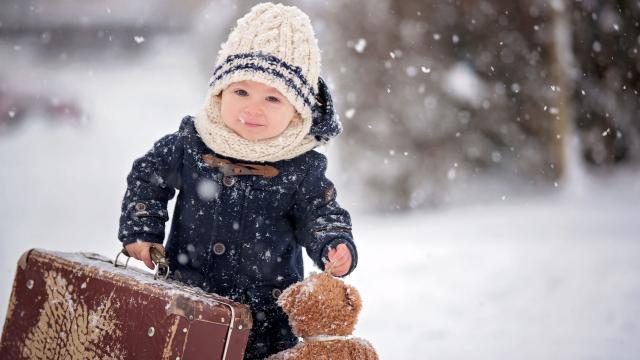 Baby playing with teddy in the snow, winter time. Little toddler boy in blue coat, holding suitcase and teddy bear, playing outdoors in winter park