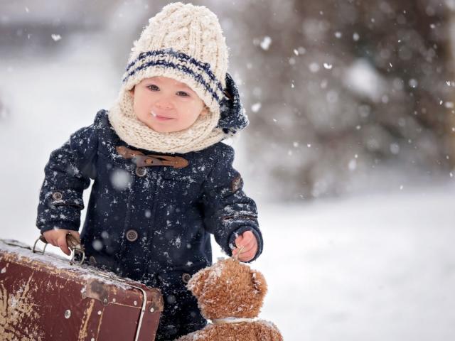 Baby playing with teddy in the snow, winter time. Little toddler boy in blue coat, holding suitcase and teddy bear, playing outdoors in winter park