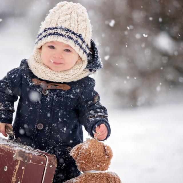 Baby playing with teddy in the snow, winter time. Little toddler boy in blue coat, holding suitcase and teddy bear, playing outdoors in winter park