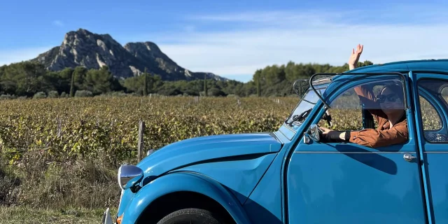 Une femme à bord d'une 2CV bleue devant un paysage de Provence