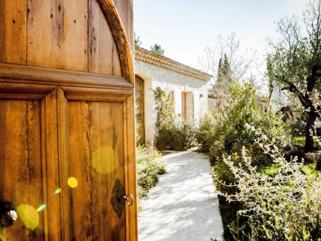 Une grande porte en bois qui s'ouvre sur l'entrée d'une bastide provençale et son jardin