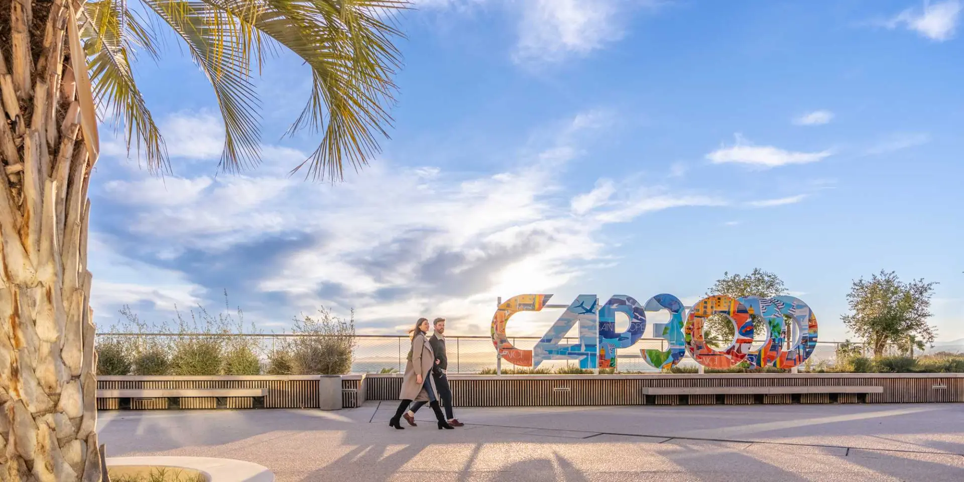 Pedestrian promenade with palm trees overlooking the sea, featuring a large colorful “CAP3000” sign and people walking along the waterfront under a bright sky.