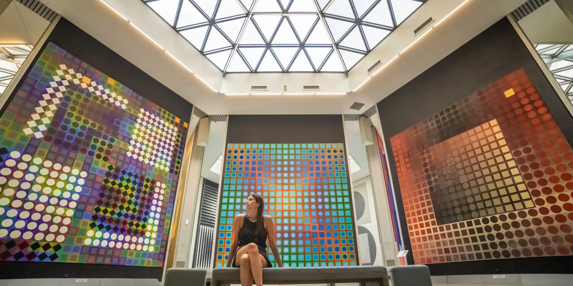 Interior of a contemporary art museum with large colorful geometric artworks displayed on the walls, a bench in the center, and a glass ceiling letting in natural light.