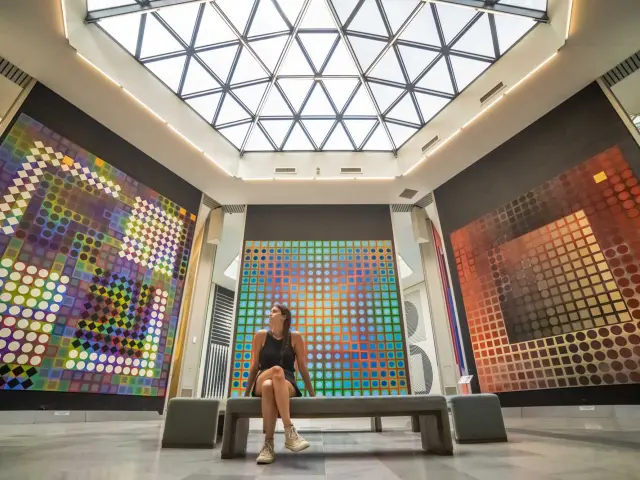 Interior of a contemporary art museum with large colorful geometric artworks displayed on the walls, a bench in the center, and a glass ceiling letting in natural light.