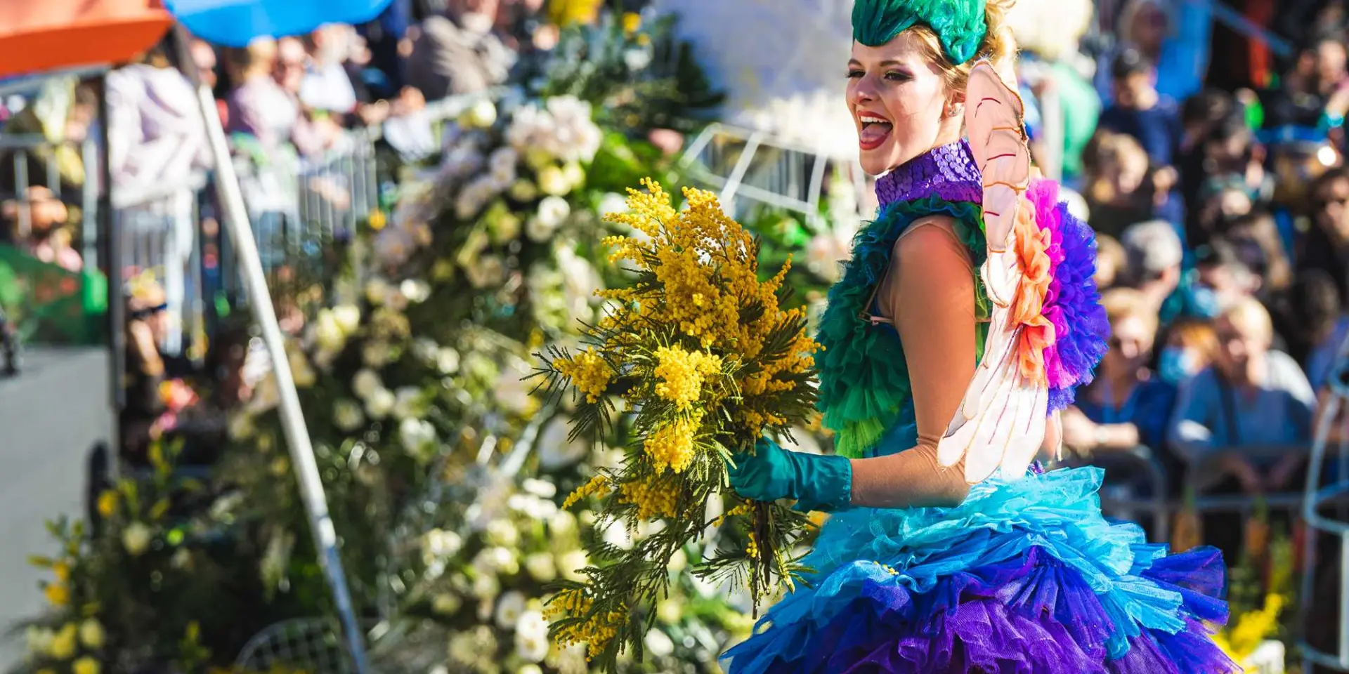 Carnaval de Nice parade scene featuring a performer in a colorful costume holding a bouquet of yellow mimosa, with floral floats and a festive crowd lining the street.