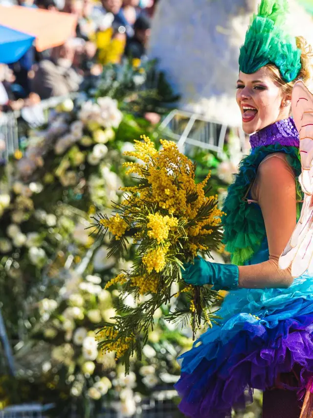 Carnaval de Nice parade scene featuring a performer in a colorful costume holding a bouquet of yellow mimosa, with floral floats and a festive crowd lining the street.