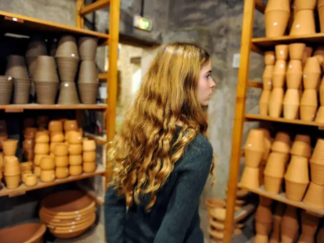Interior of the Poterie Ravel workshop, with shelves filled with terracotta pots and bowls, and a visitor walking between rows of handcrafted ceramic pieces.