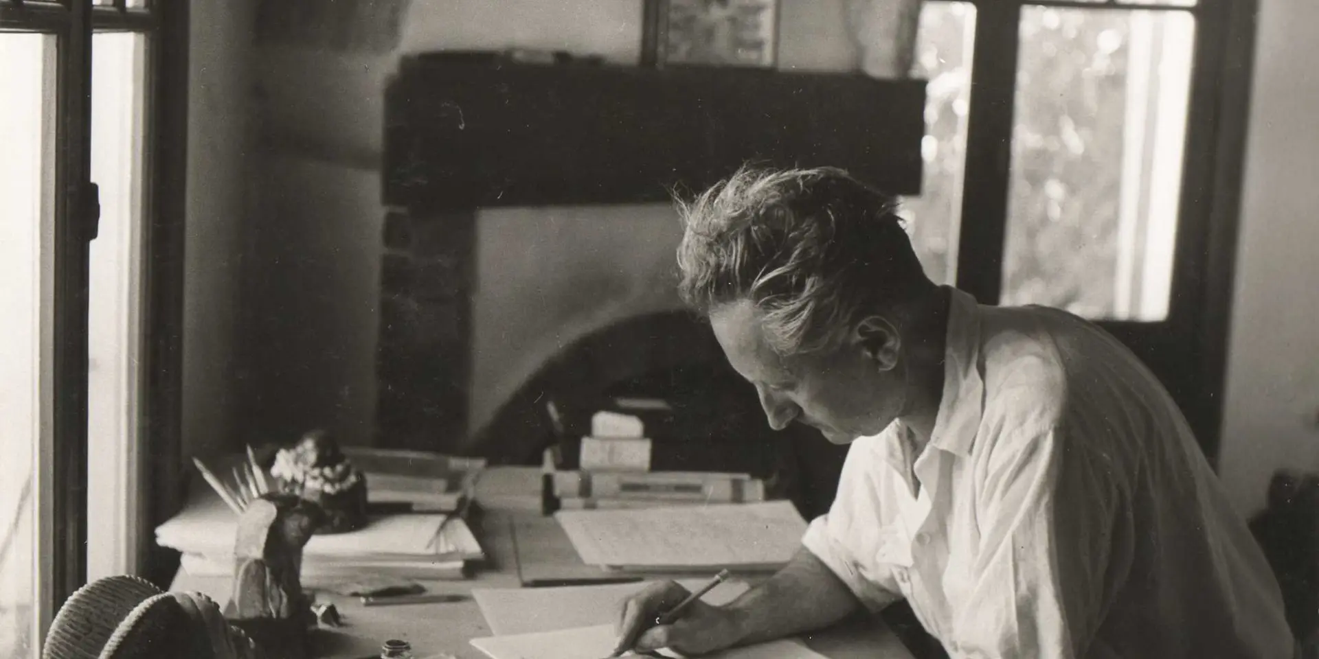 Writer Jean Giono seated at a wooden desk, writing by hand among notebooks, books, and papers in a light-filled interior with a fireplace and large windows.