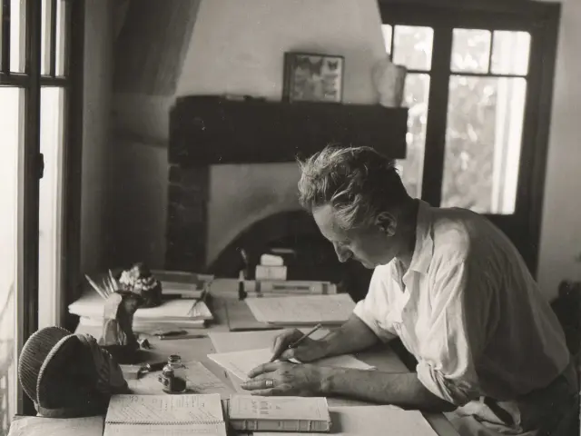 Writer Jean Giono seated at a wooden desk, writing by hand among notebooks, books, and papers in a light-filled interior with a fireplace and large windows.
